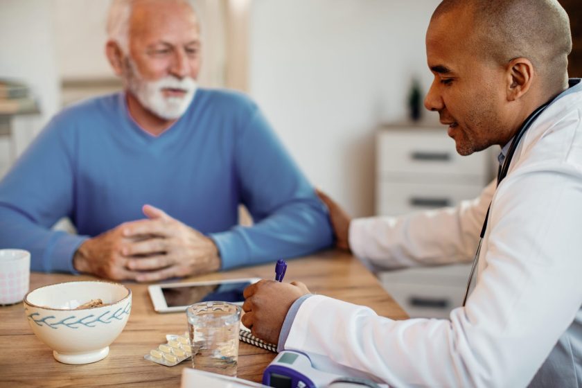 Black general practitioner writing notes while communicating with mature man during home visit.