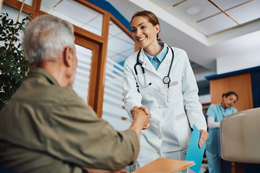 Happy female doctor and mature patient shaking hands while greeting in waiting room at medical clinic.