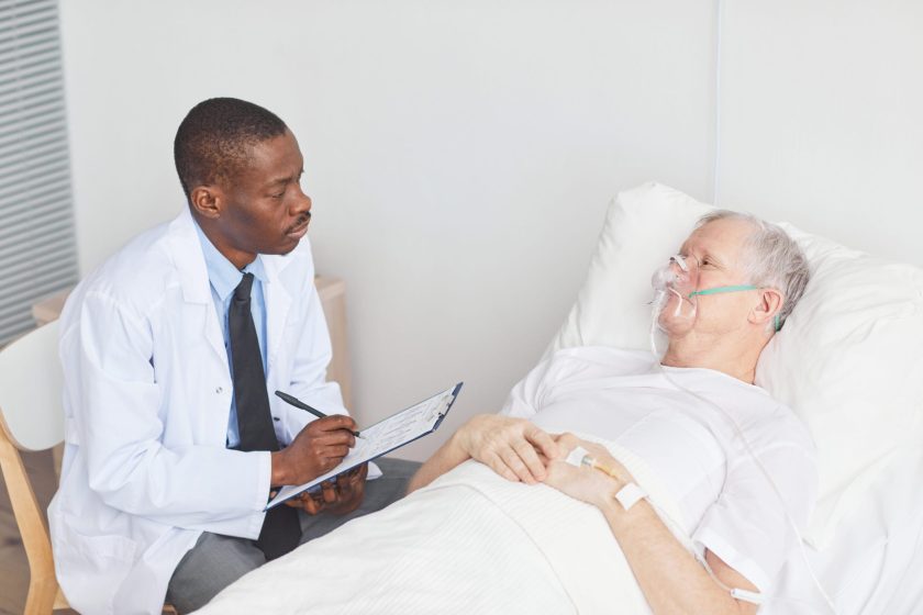 Portrait of African-American doctor talking to senior man lying in hospital bed with oxygen supplementation mask, copy space